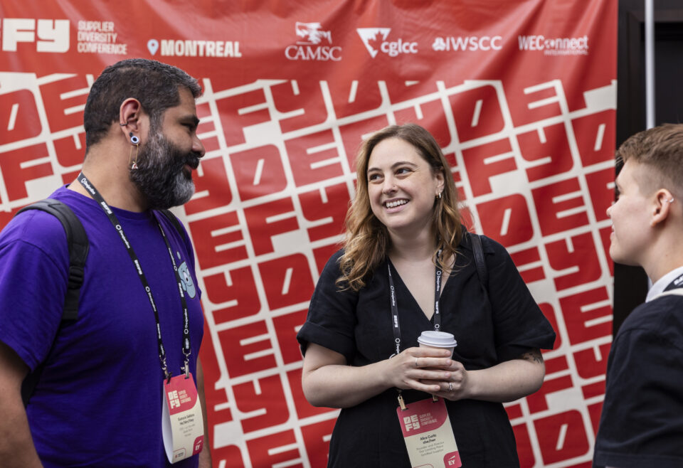 Three people standing and talking in front of a large sign with DEFY branding in red text.